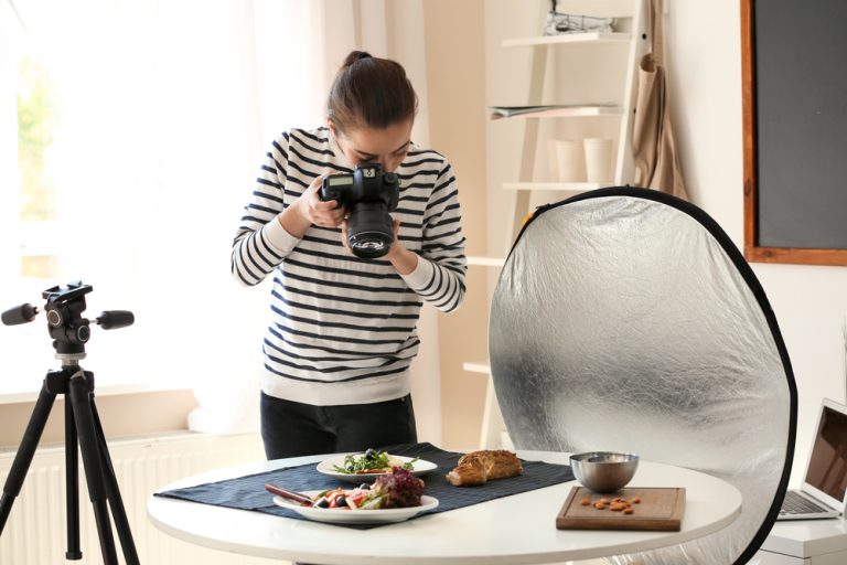 Woman photographing food on a table with a camera, using a reflector and tripod nearby in a well-lit room for her food photography blog.