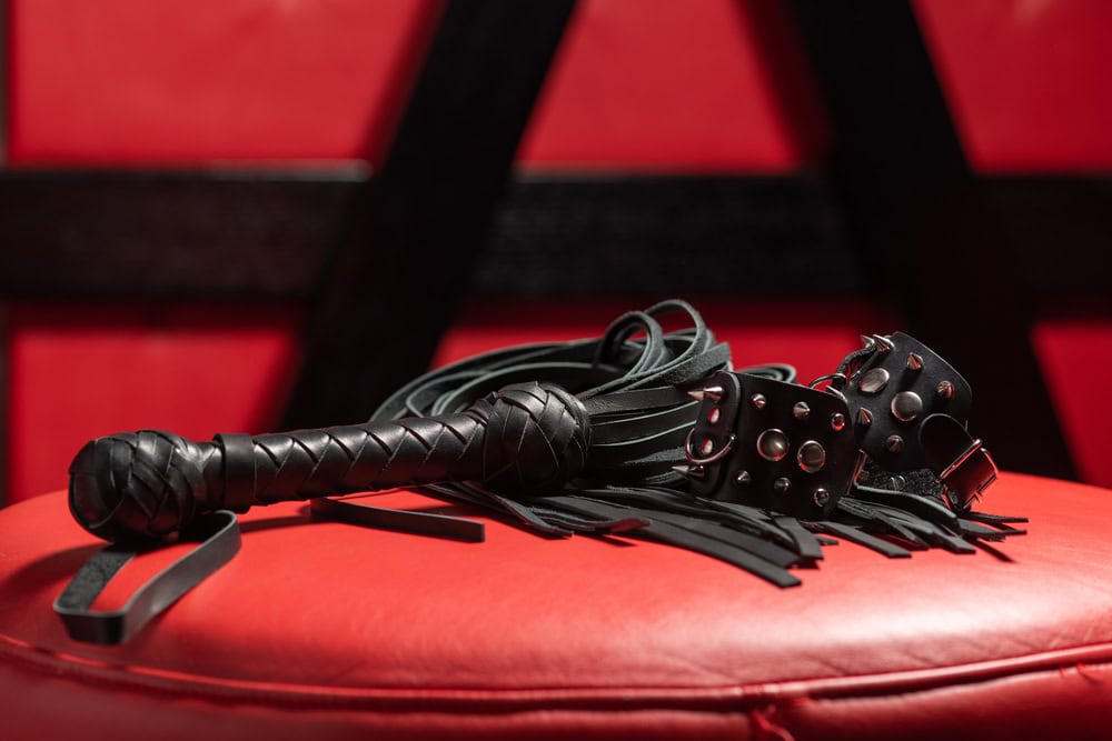 A black leather flogger and studded black wrist cuffs rest on a red padded surface, setting the scene for exploring emotional safety kink, with dark and red furniture visible in the background.